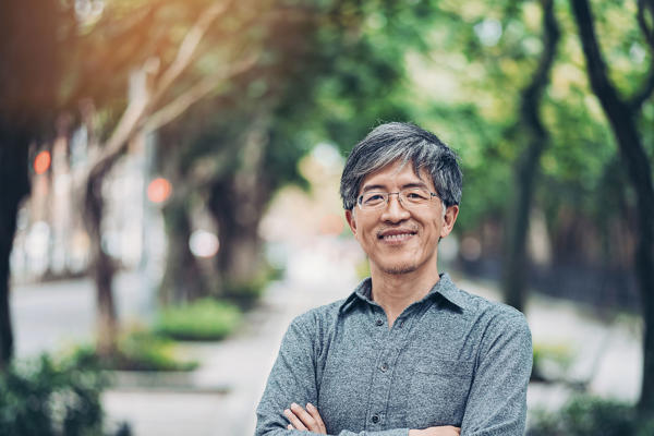 An older man poses standing outside while smiling.