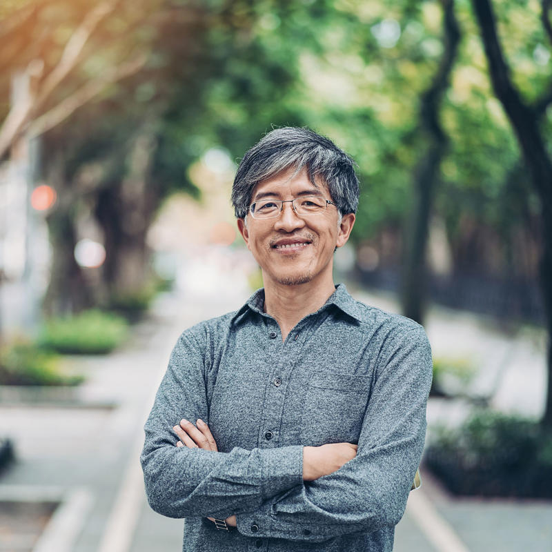 An older man poses standing outside while smiling.