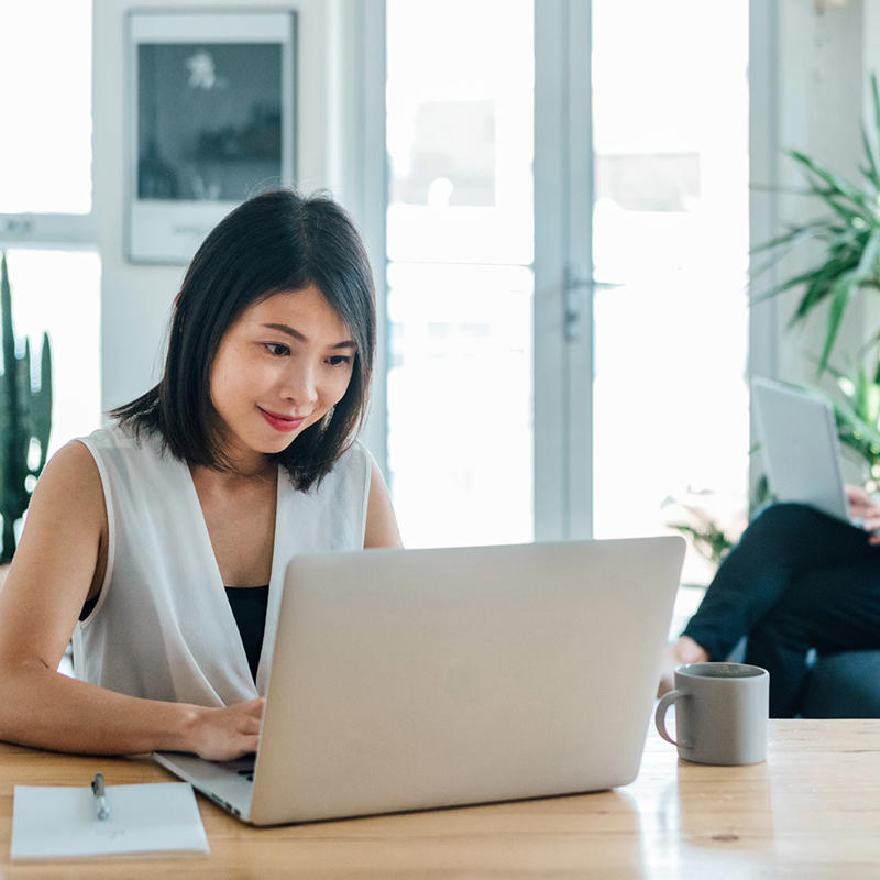 A woman works on a laptop while a man works on his laptop in the background.