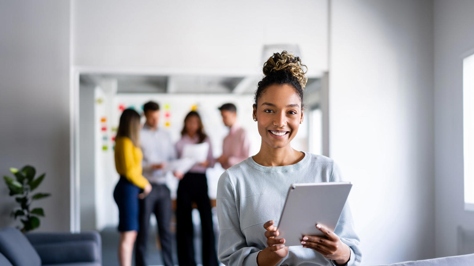 A woman stands with a tablet with coworkers in the background.