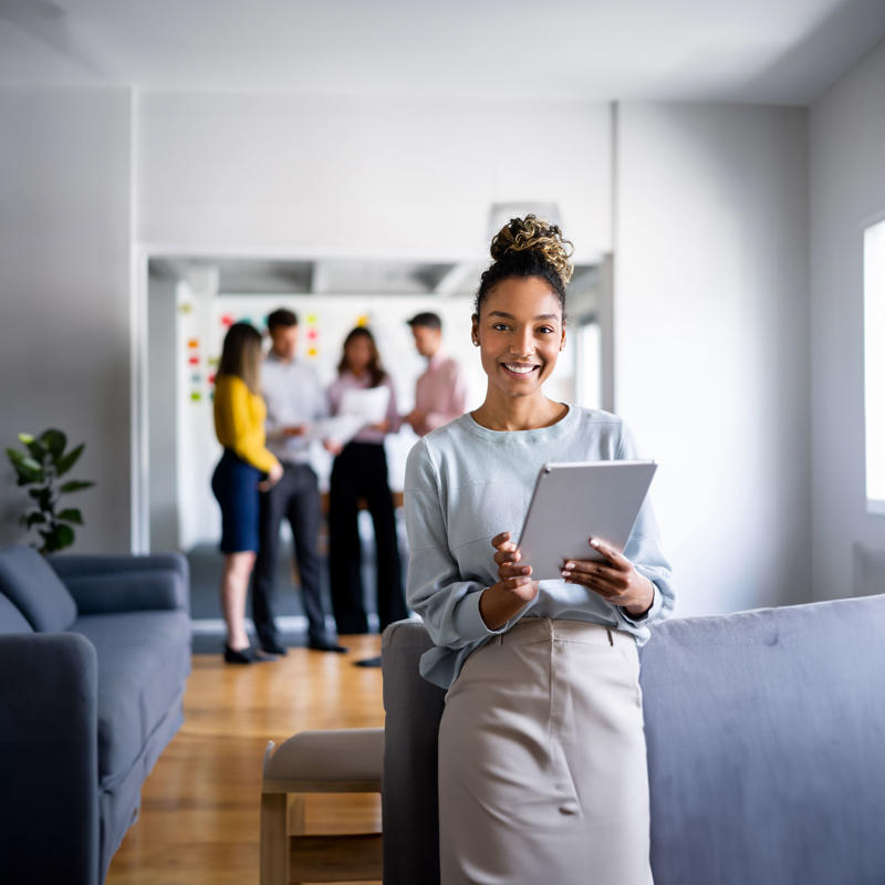A woman stands with a tablet with coworkers in the background.